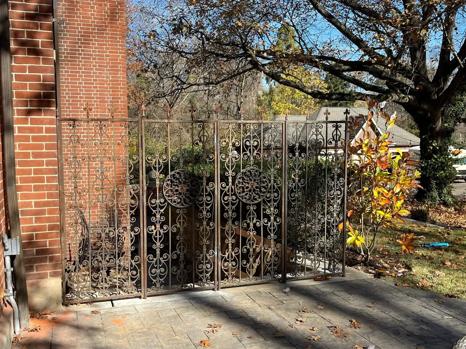 Brown wrought iron fence in front of a brick building, with trees and sunlight in the background.