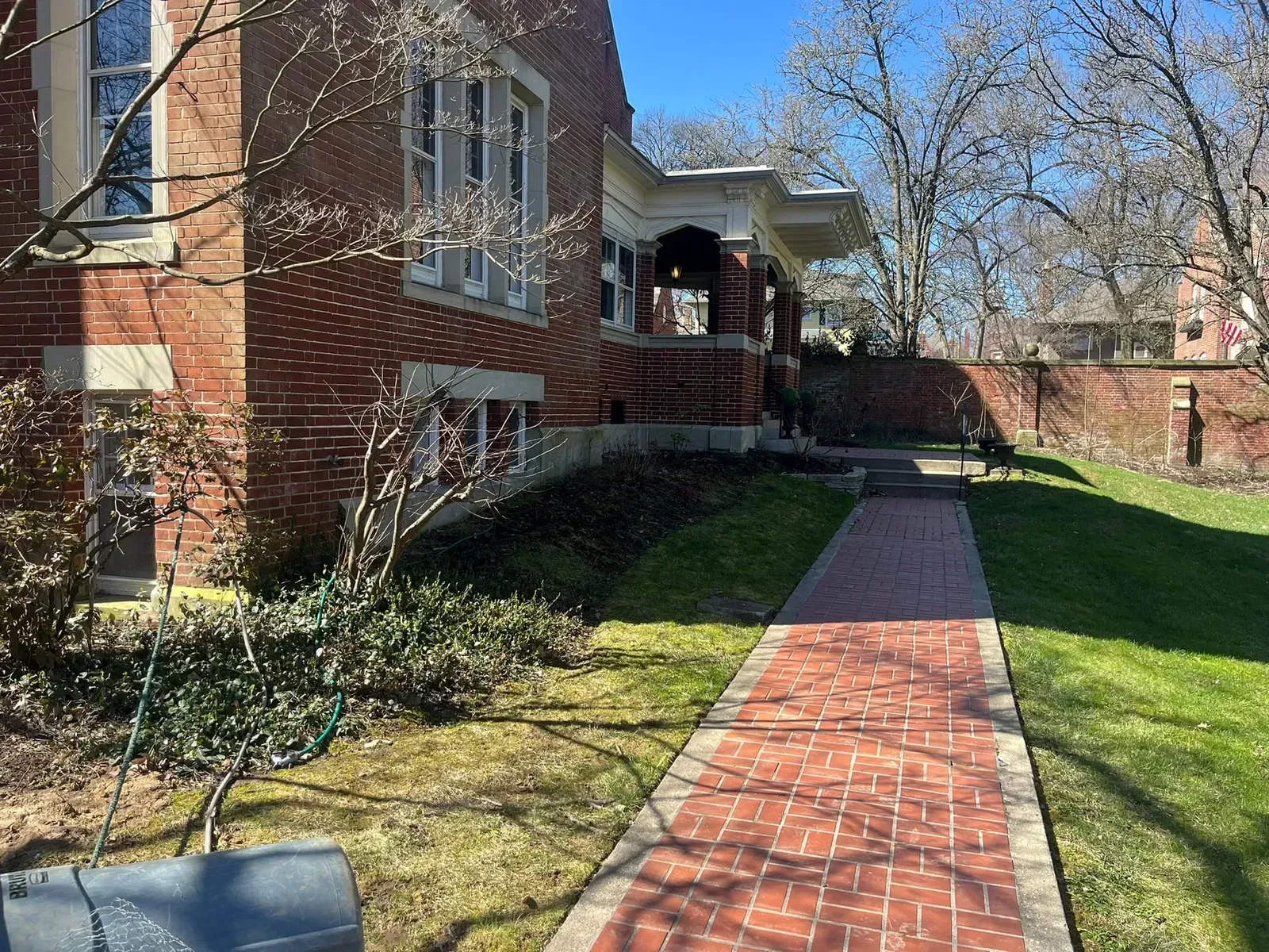 Brick pathway leading to a brick house with a porch. Green grass and trees. Bright, sunny day.
