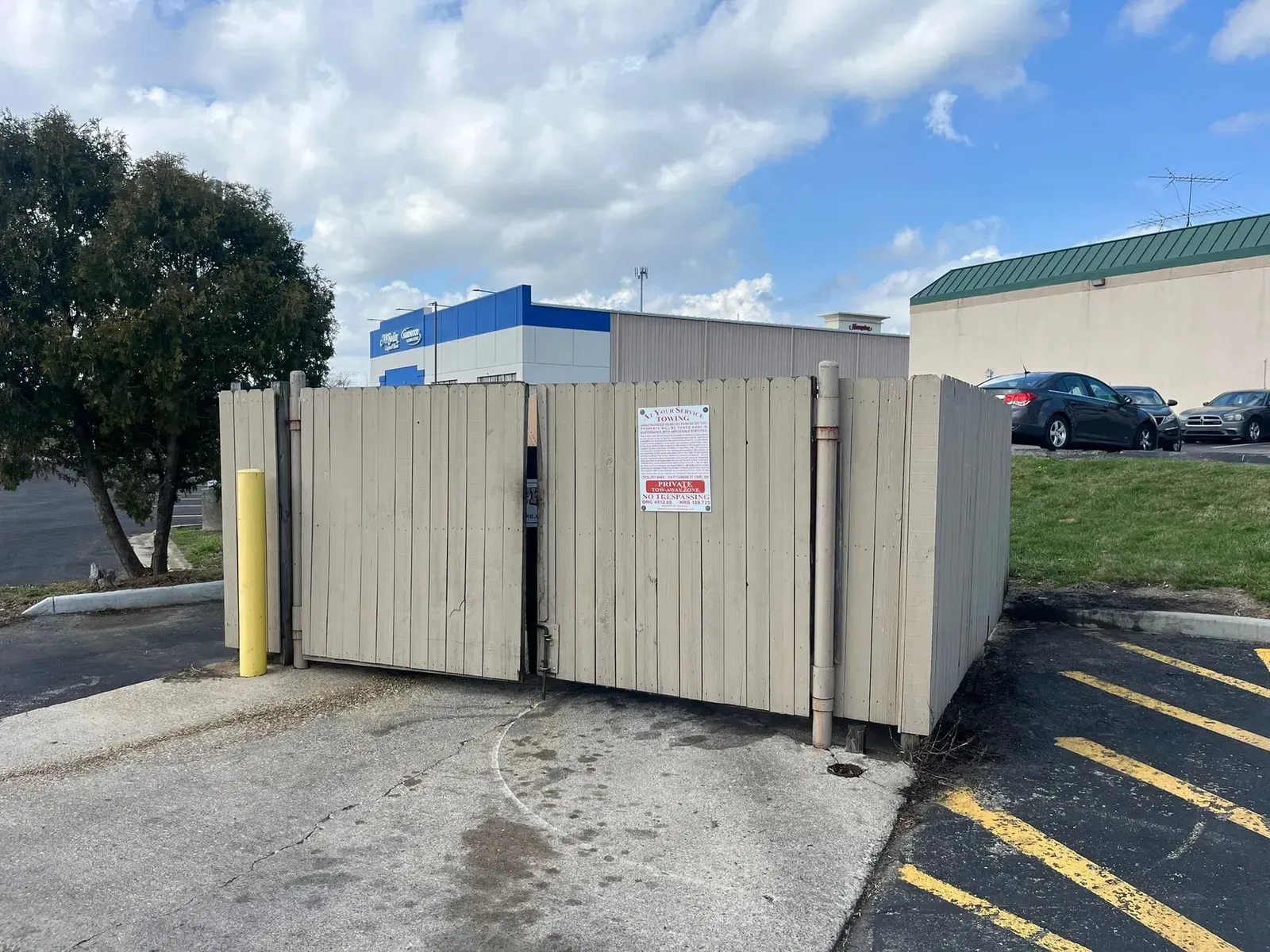 Dumpster enclosure with closed, weathered, tan gates in a parking lot. Buildings and cars in background.