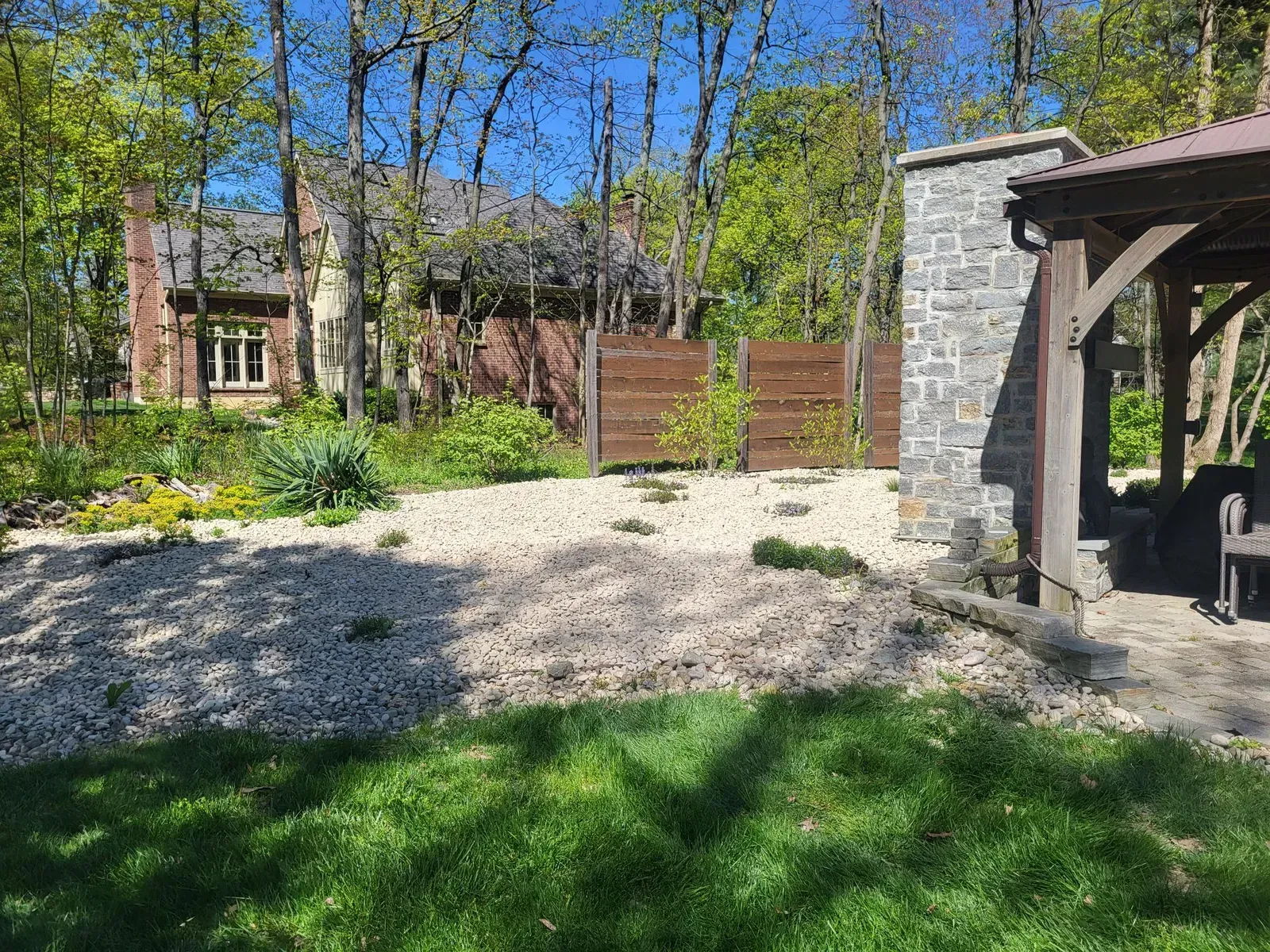 Backyard scene with gravel bed, wooden fence, gazebo, and a house with trees.