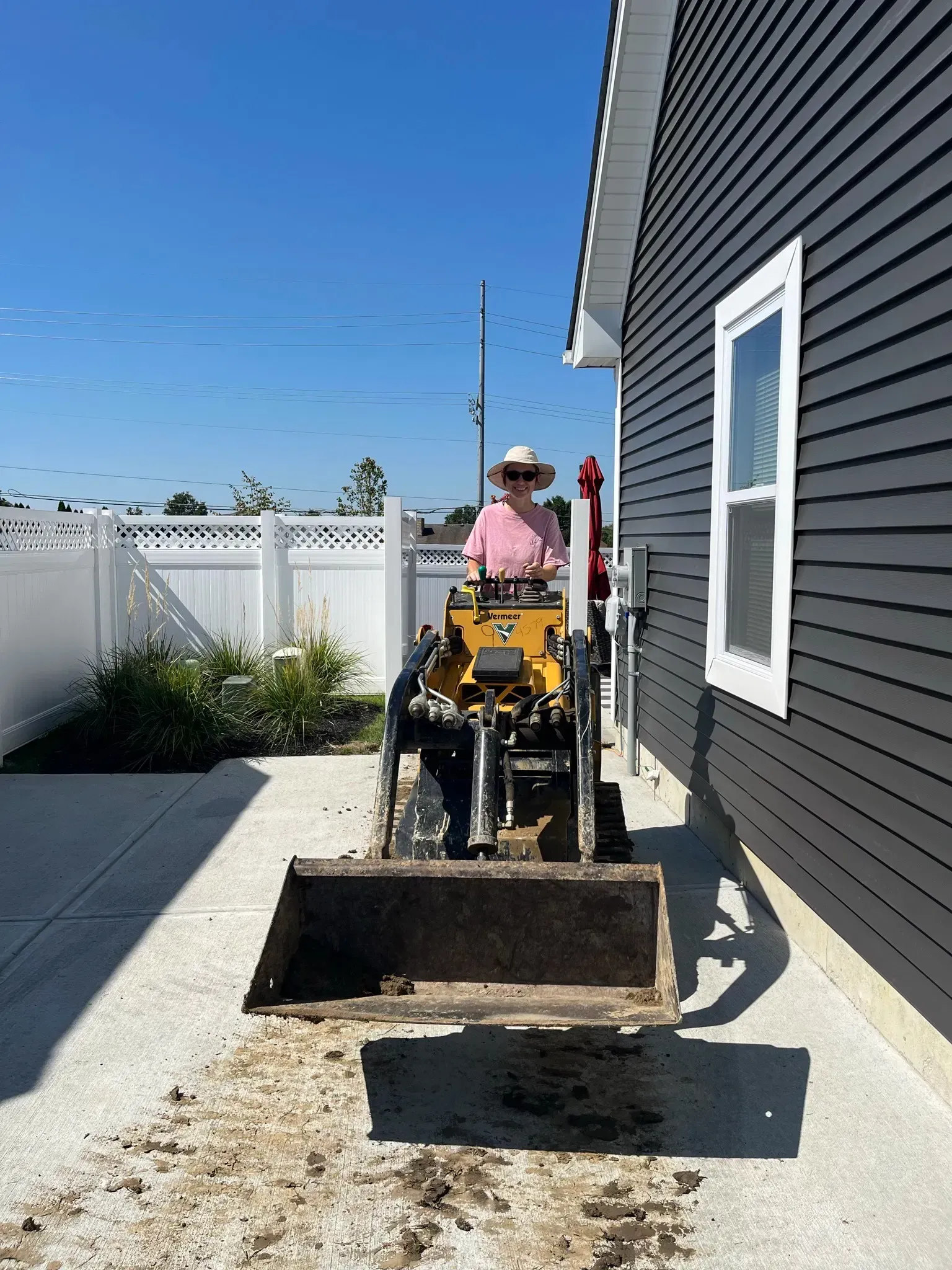 Person operating small excavator near a dark house with a white window, and a fence under a blue sky.