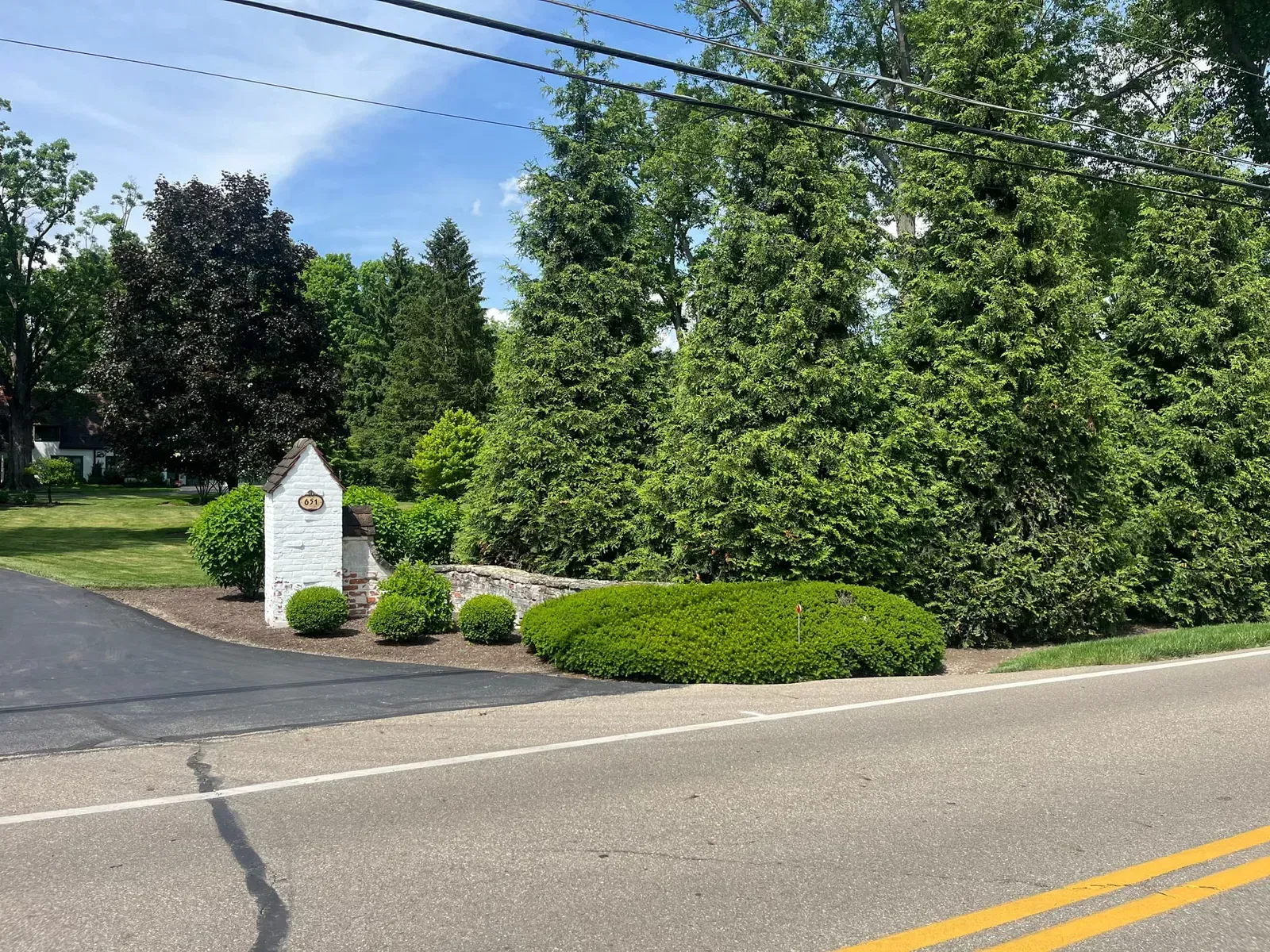 Stone entrance with a sign, surrounded by trees and bushes, next to a road.