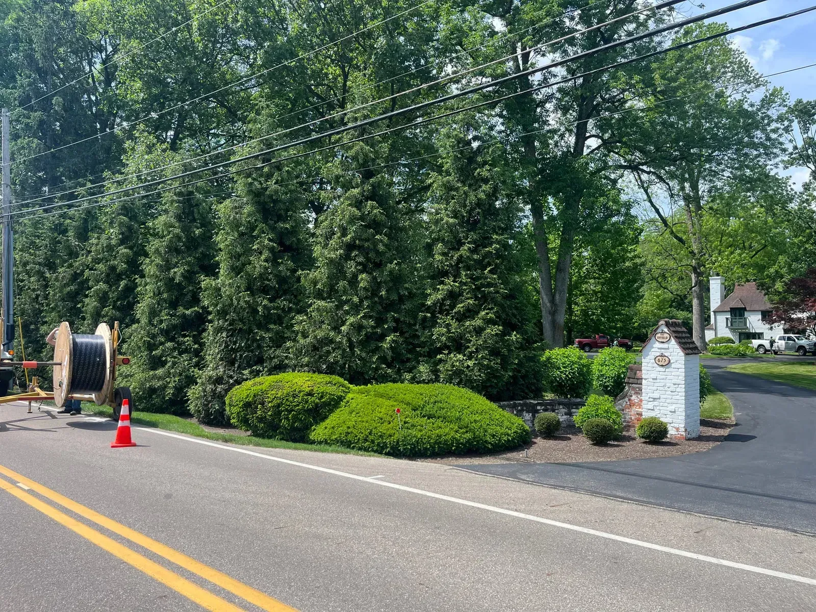 A road with telephone wires above, with a spool of cable on the side. Trees and shrubbery frame the view.