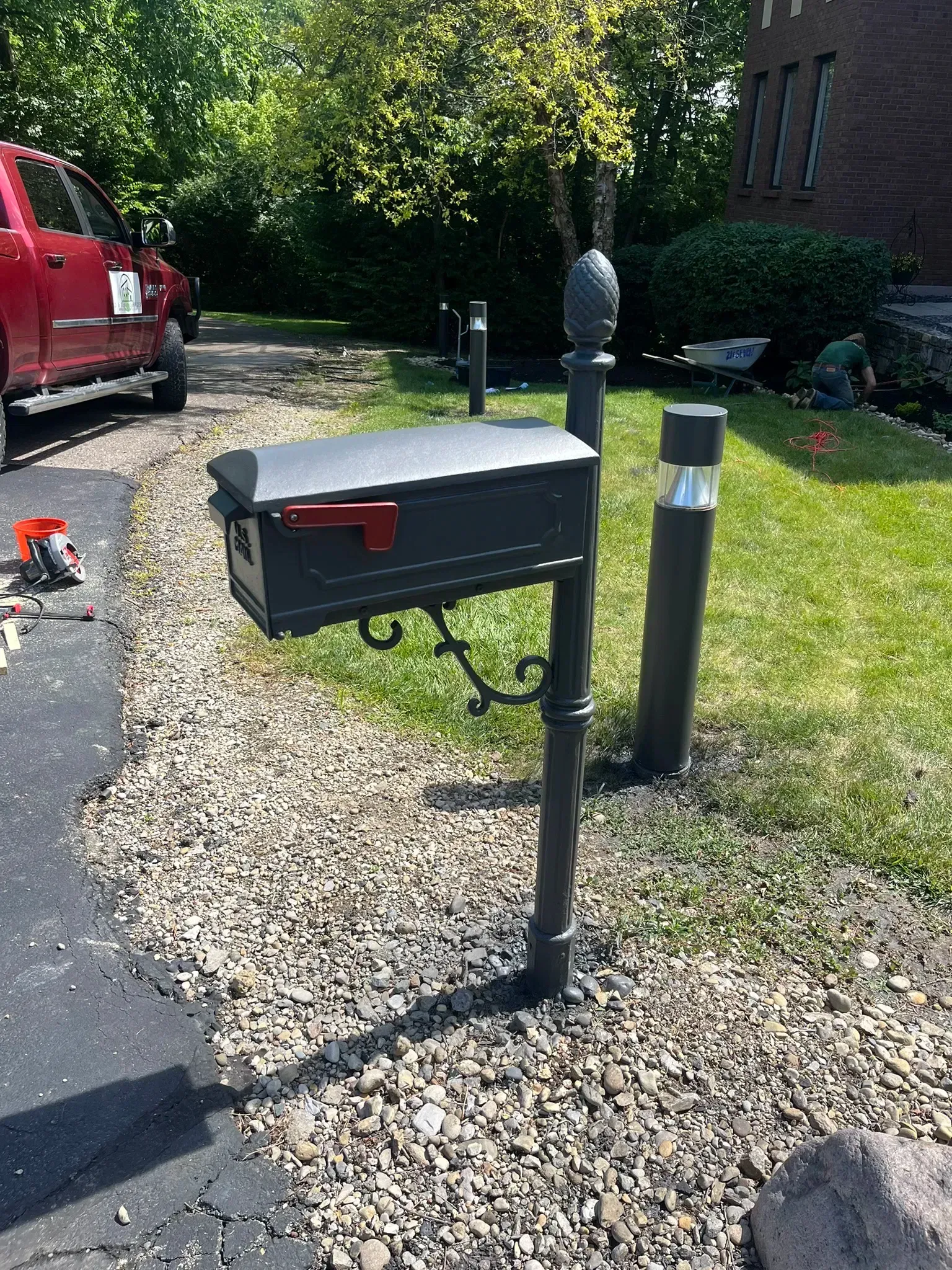 Black mailbox on ornate post with gravel ground cover, red truck in background.