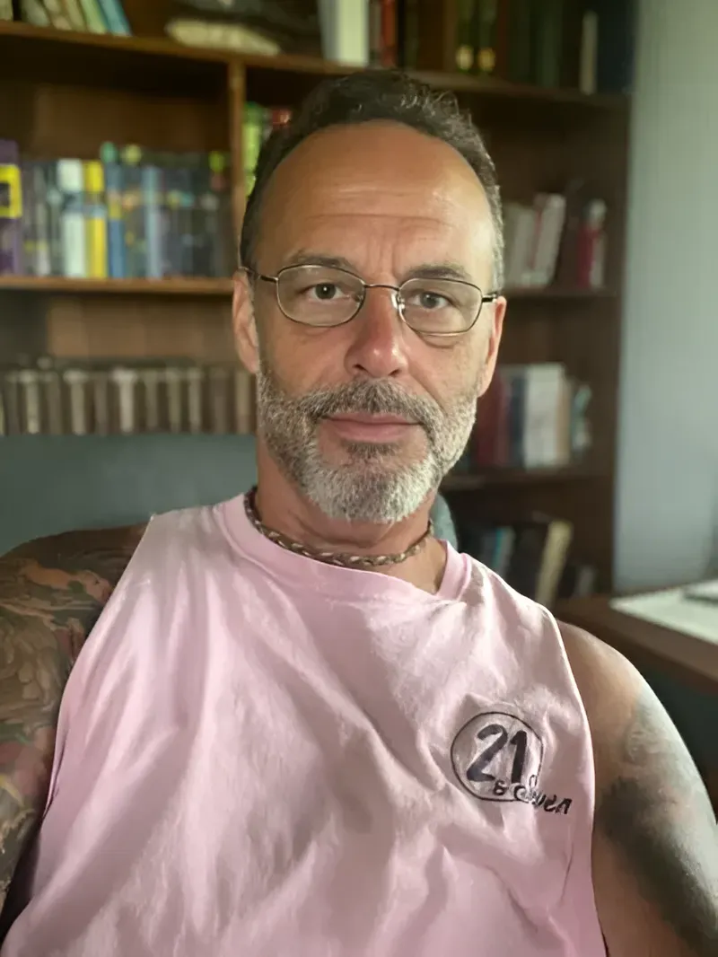 Man with glasses, graying hair and beard, pink tank top, in a room with bookshelves.