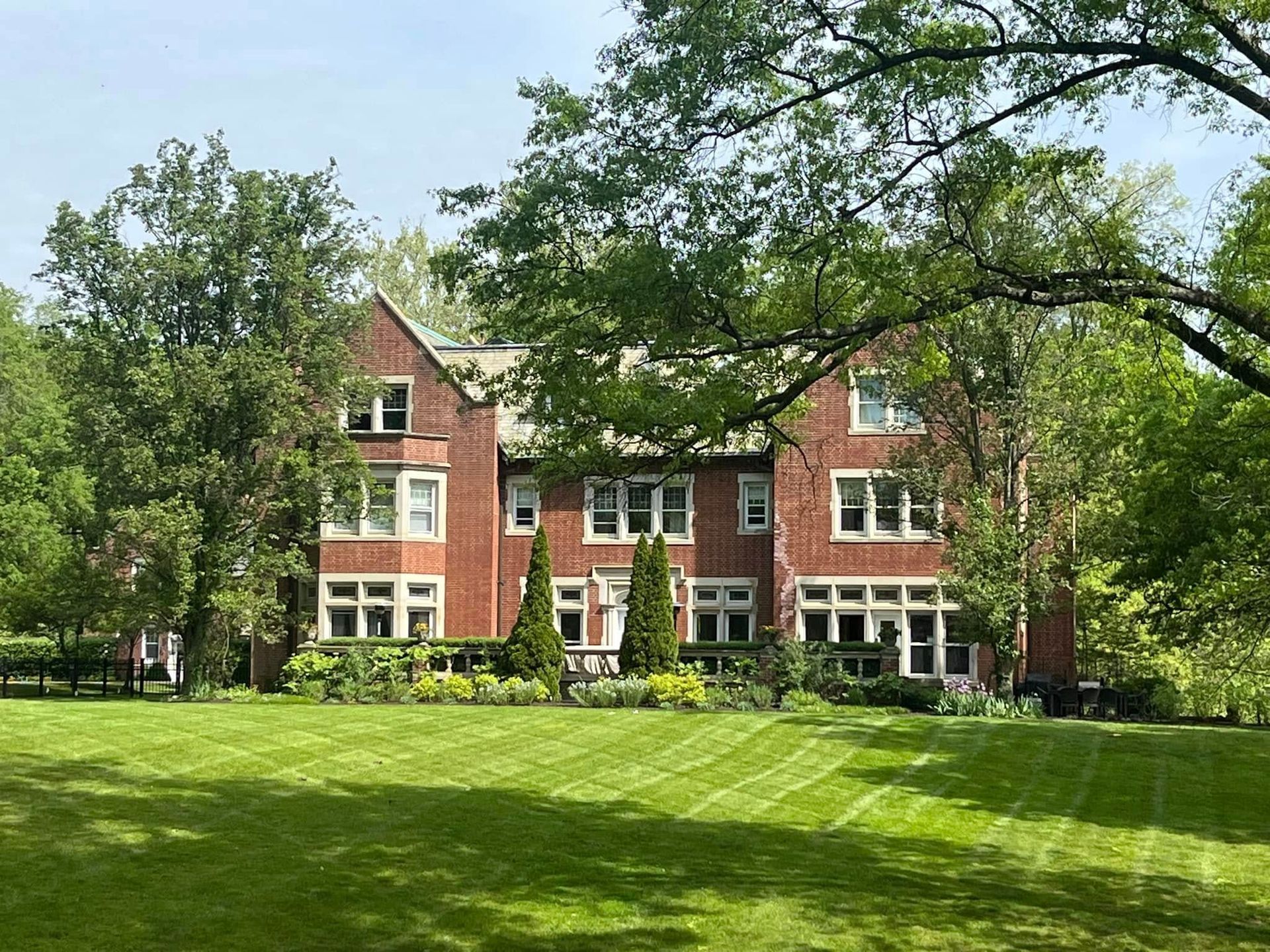 Red brick mansion nestled in green lawn, surrounded by trees. Sunny day.