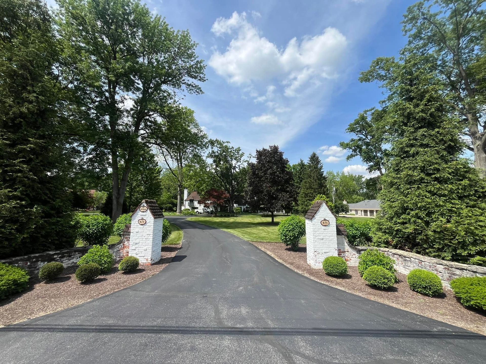Paved driveway leading to a white house, flanked by brick pillars and greenery under a blue sky.