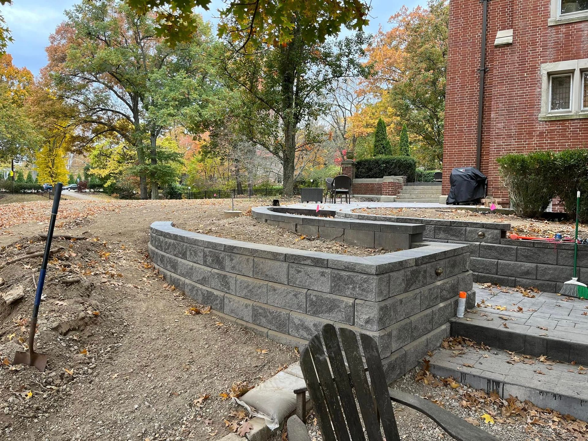 A tiered retaining wall of gray blocks next to a brick house and trees. Fall foliage, gravel ground.