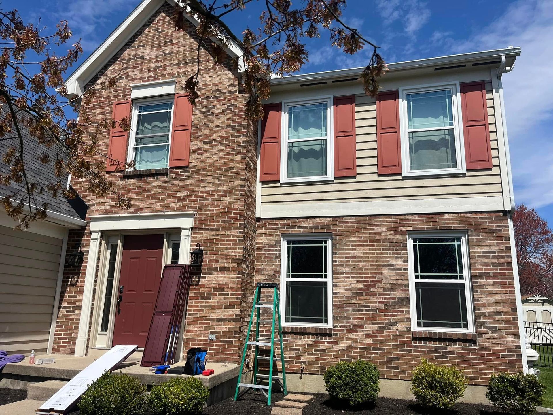 Two-story brick house with red shutters. A ladder and materials are on the ground; cloudy blue sky.