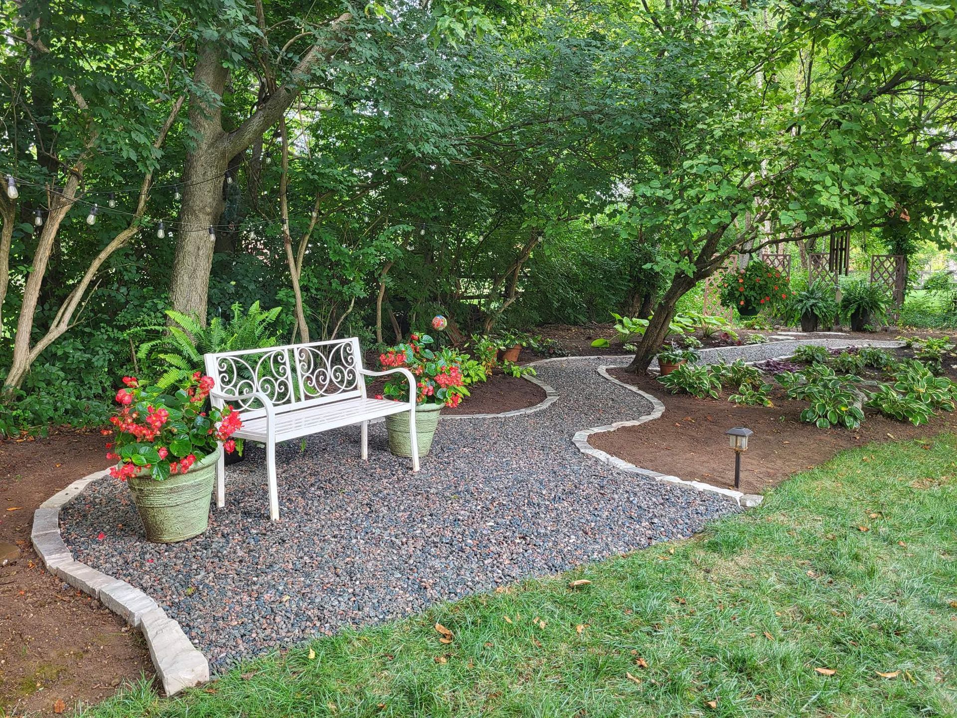 White bench sits on a gravel path lined with plants, beneath trees in a garden.