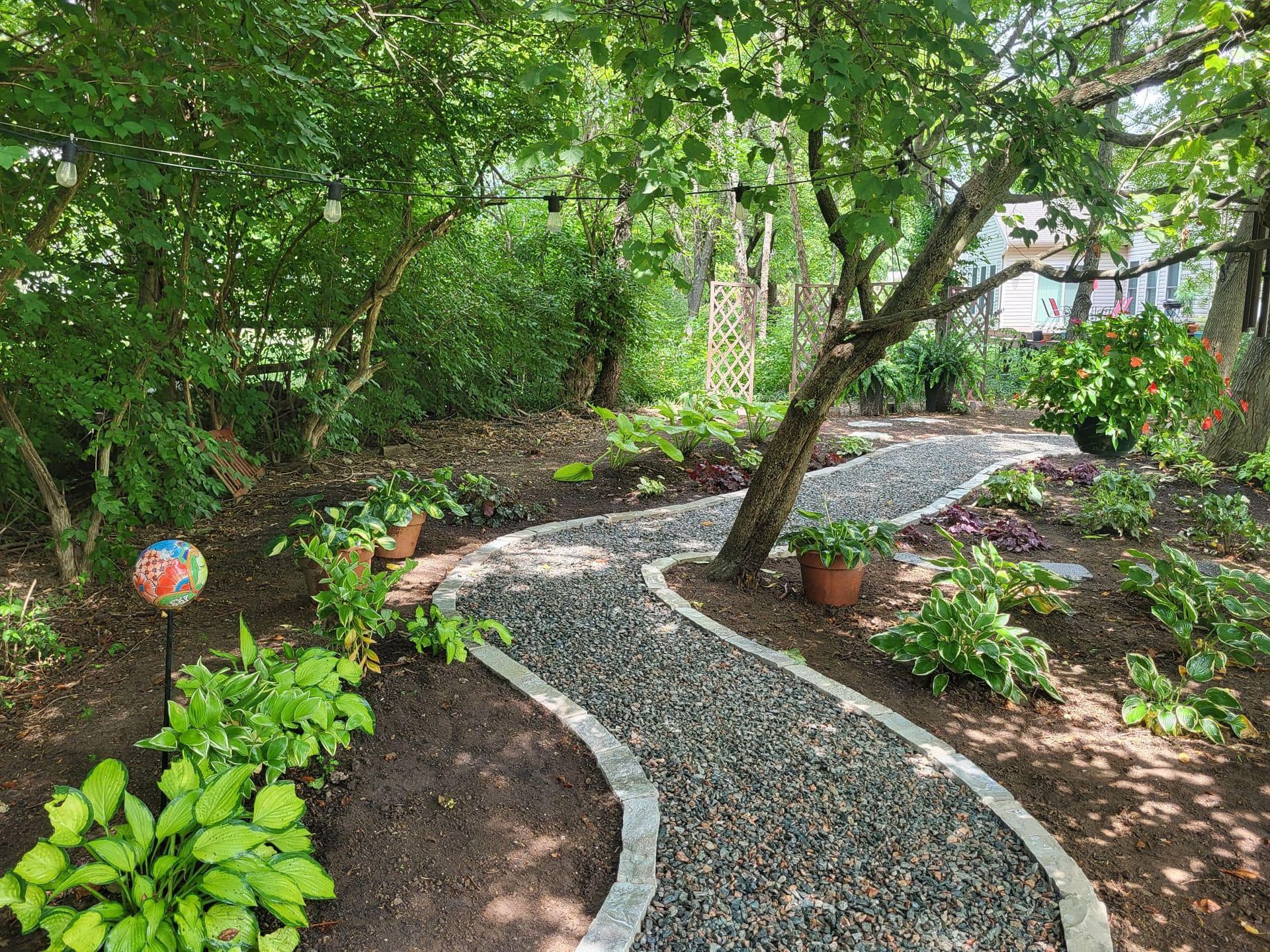 Winding pebble path through a lush garden with potted plants and trees on a sunny day.