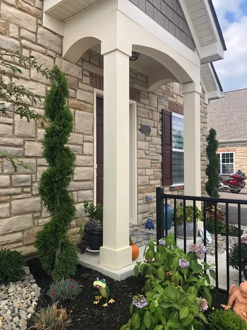 Stone house entrance with columns, spiral tree, and garden plants.