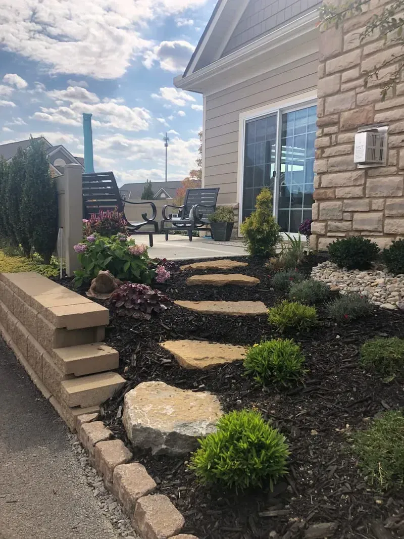 Stone steps lead to a patio with chairs, next to a house with a stone facade and landscaping.