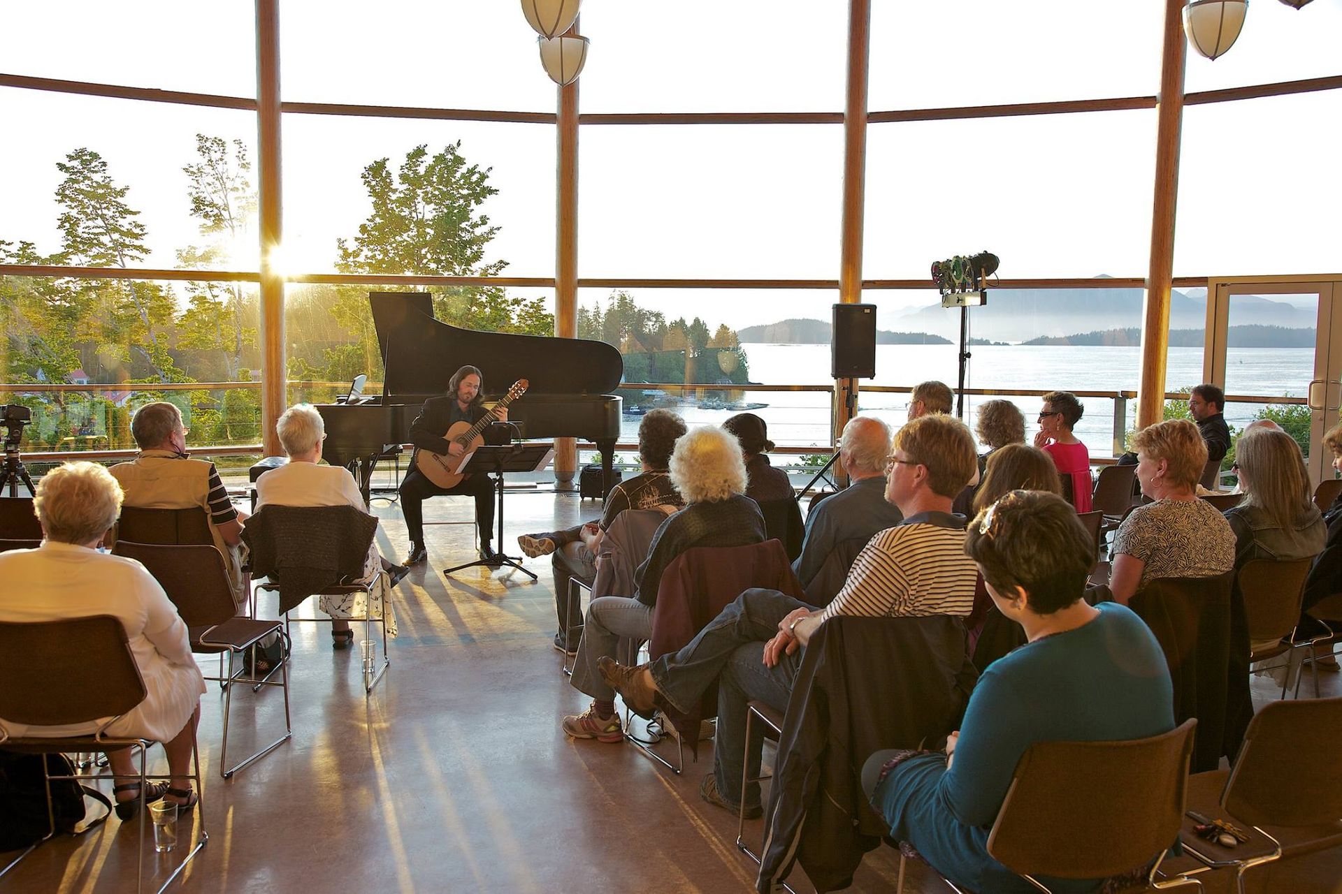 A group of people are sitting in front of a piano while a man plays a guitar.