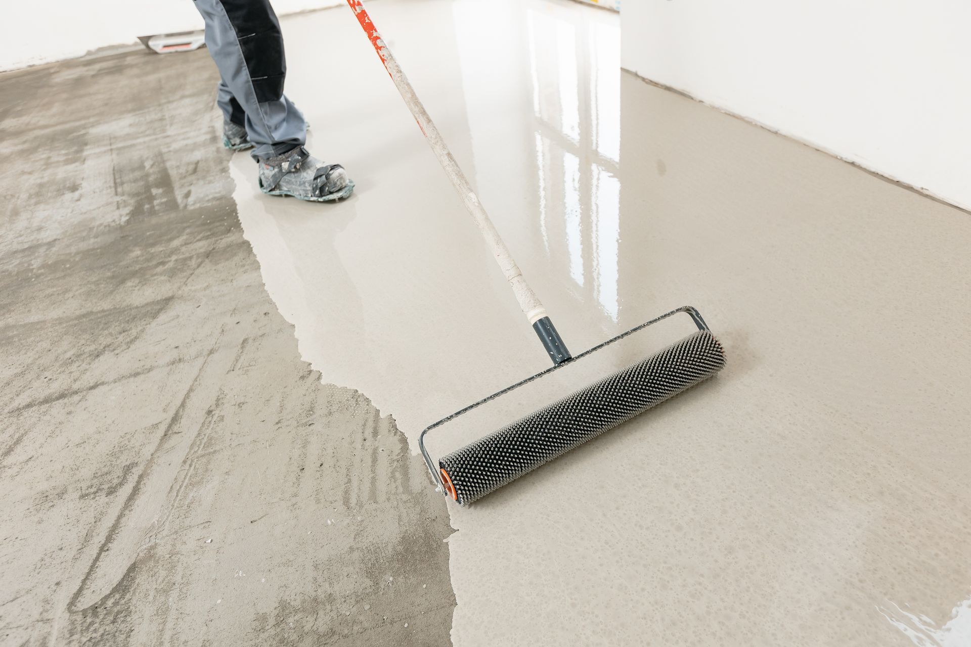 Person applying light gray sealant to a floor with a roller applicator in a room.