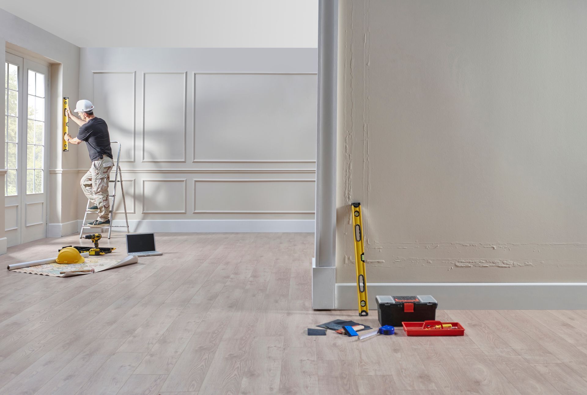 Person on ladder levels a wall in a room during renovation, with tools nearby.