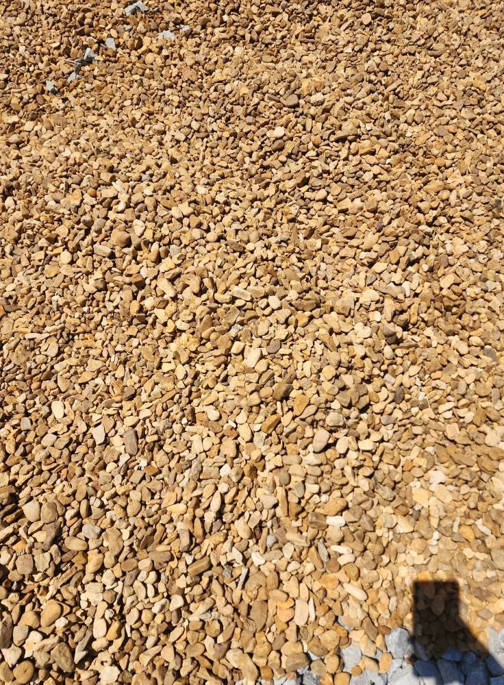 Gravel ground covering, tan and brown pebbles. Shadow of a person visible in lower right corner.