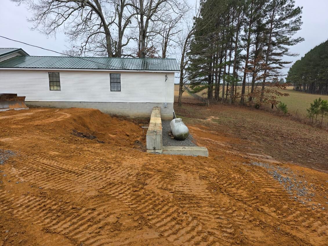 A propane tank beside a white building on a graded, muddy lot with tire tracks, trees in the background.