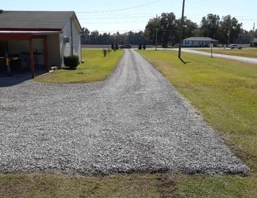 Gravel driveway extending from a small beige building to a road, surrounded by grass and blue sky.