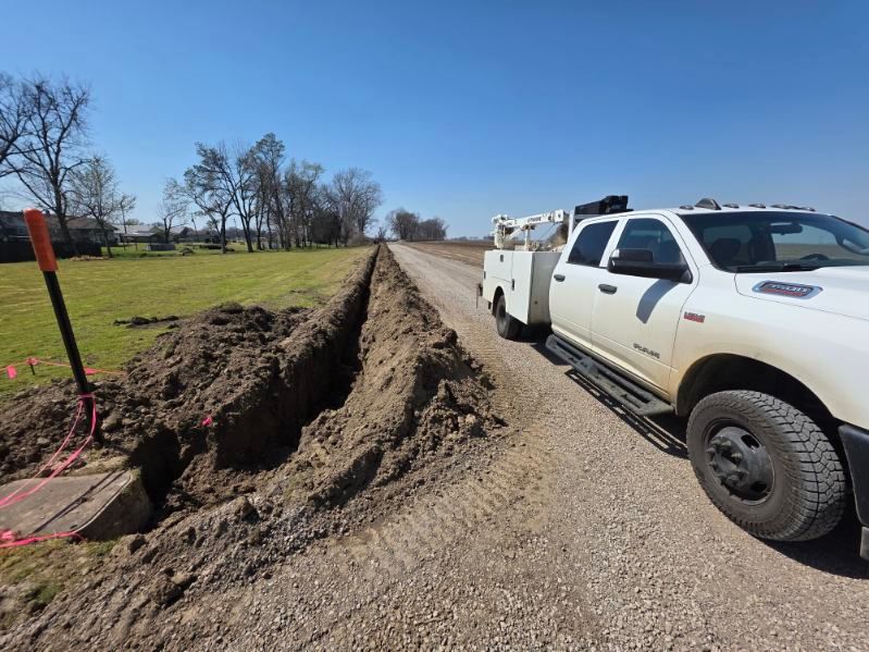White utility truck parked beside a gravel road next to a long dirt trench in a rural field