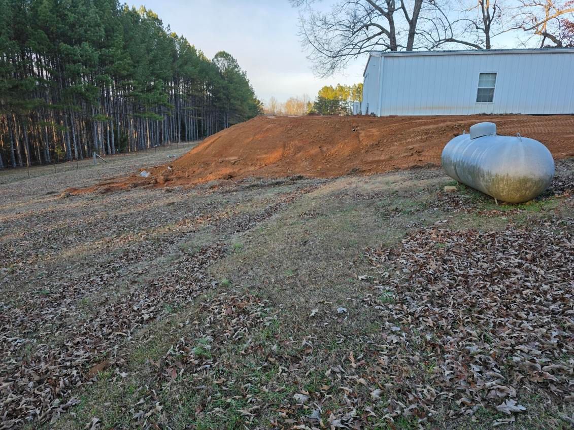 A metal propane tank next to a dirt mound, a mobile home, and trees on a grassy hill.