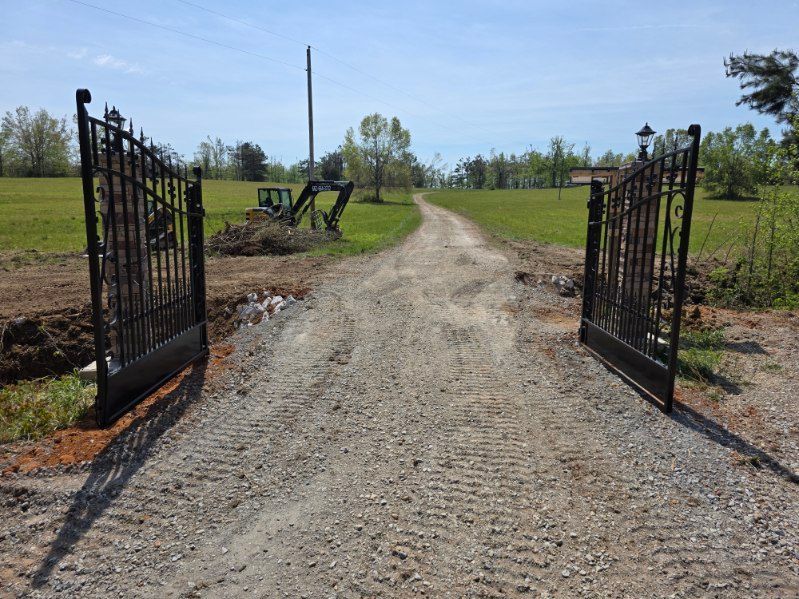 Open iron gates on a gravel driveway leading to a grassy field under a clear sky