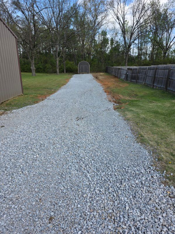 Gravel driveway leading to a metal gate, with trees and a fence on both sides.