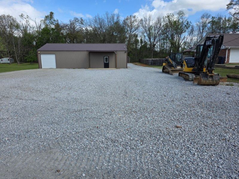 Gravel lot beside a tan building with a white garage door and construction equipment under a cloudy sky