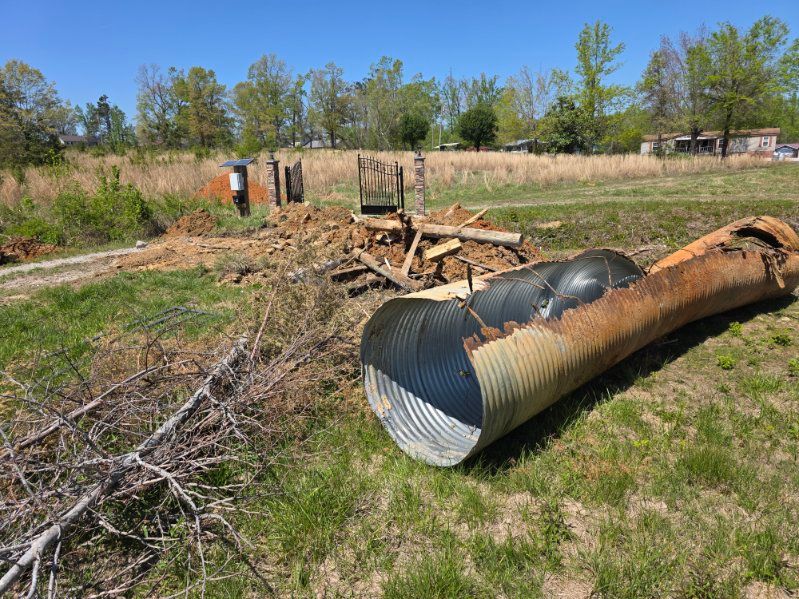 Collapsed metal culvert pipe on grassy field with scattered debris and trees in the background