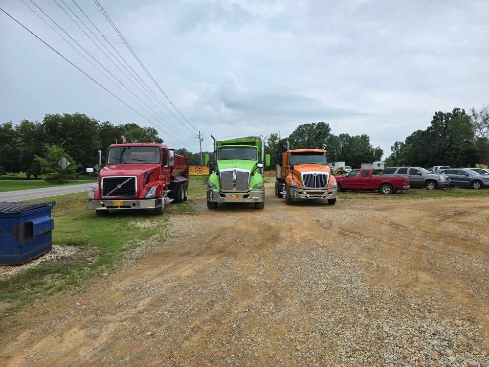 Three dump trucks, red, green, and orange, parked on gravel in front of cars and trees under a cloudy sky.