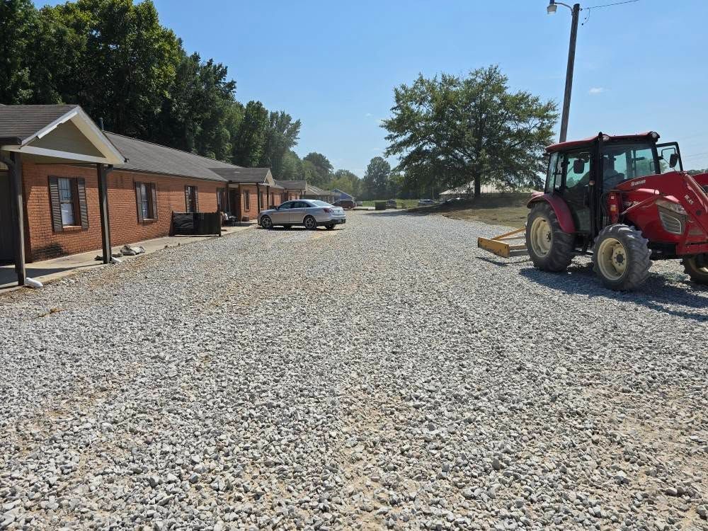 A gravel parking lot with a red tractor on the right, brick buildings on the left, a car in the distance.