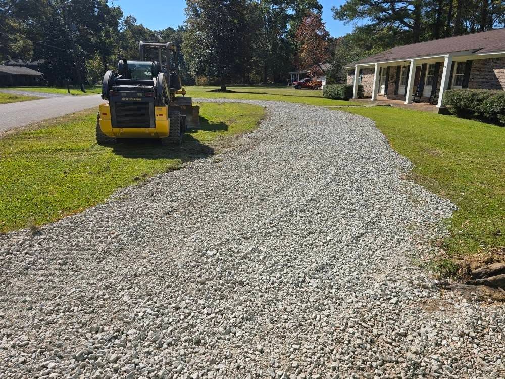 Gravel driveway in front of a house, with a yellow skid-steer loader on the left and green grass on either side.