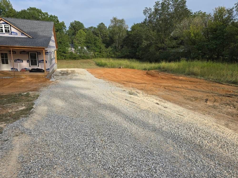 Gravel driveway leading to a partially built house on a grassy lot, trees in the background, overcast sky.