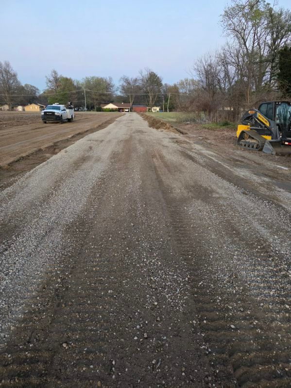 Gravel road under construction with a truck on the left and an excavator on the right, trees lining the sides