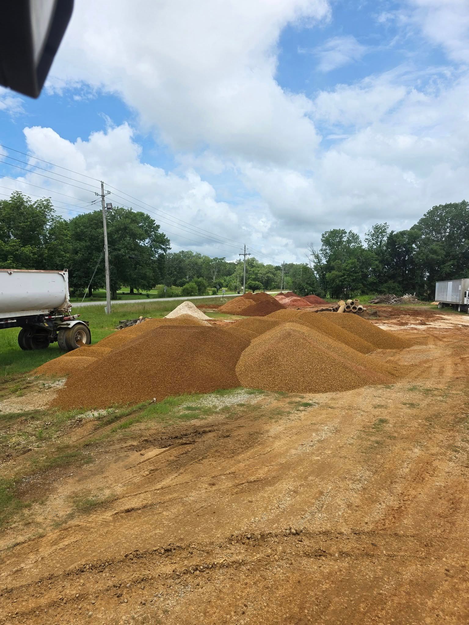Piles of gravel on dirt, with a truck, trees, and cloudy sky in the background.