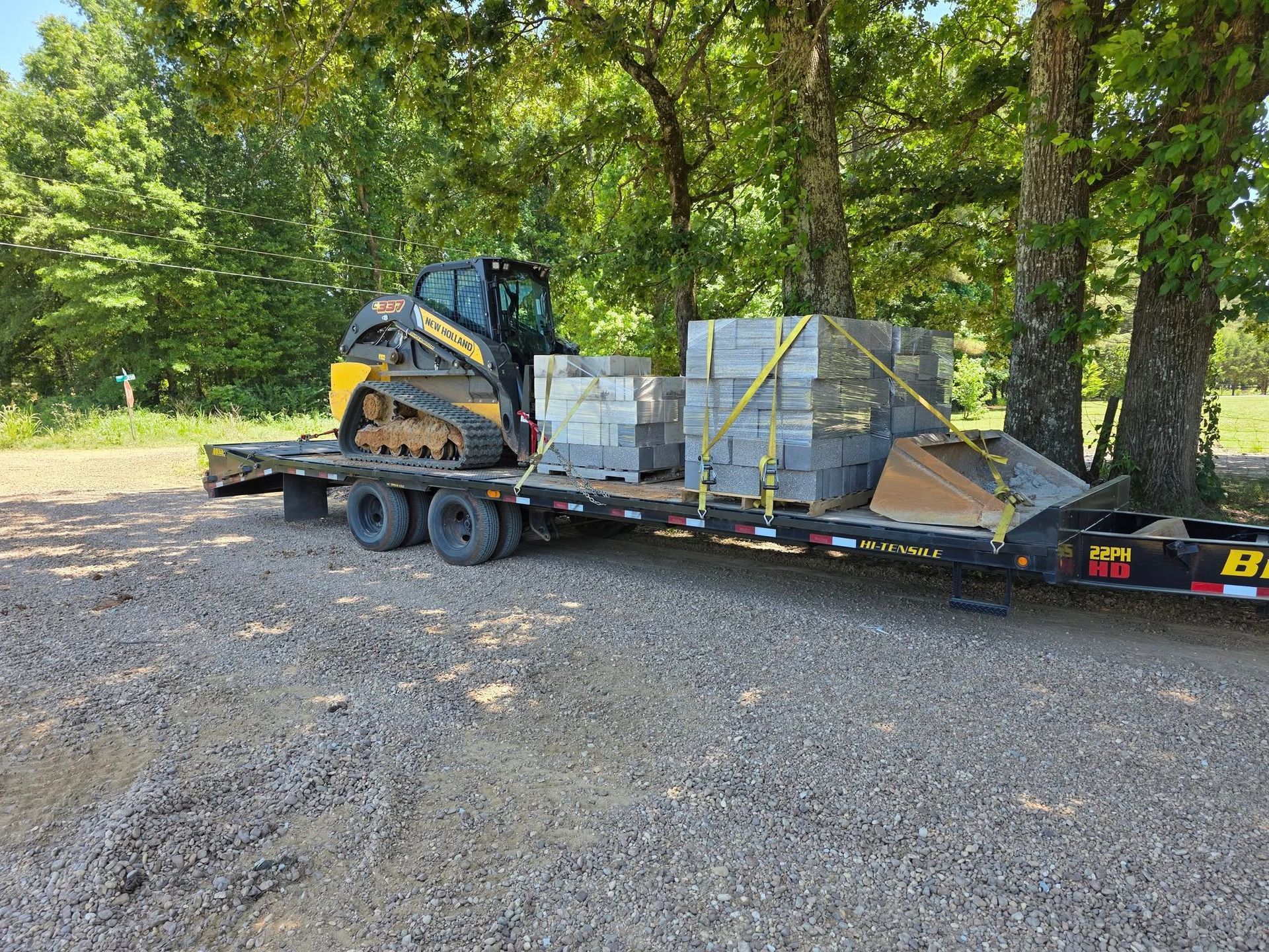 Skid steer loader and cinder blocks secured on a trailer. Trees in the background.