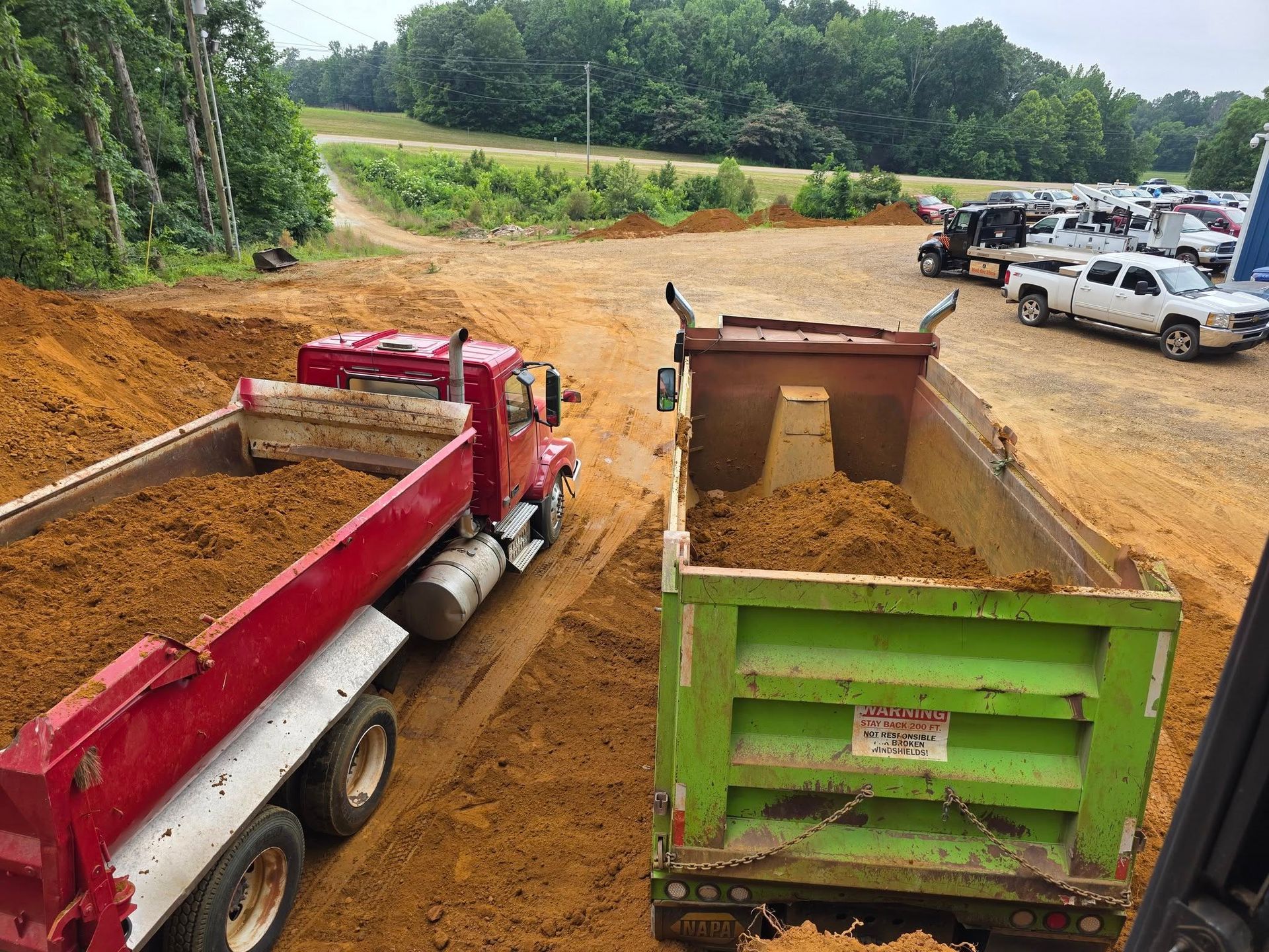 Two dump trucks loaded with dirt, one red, one green, in a construction site; a parking area with cars in the background.