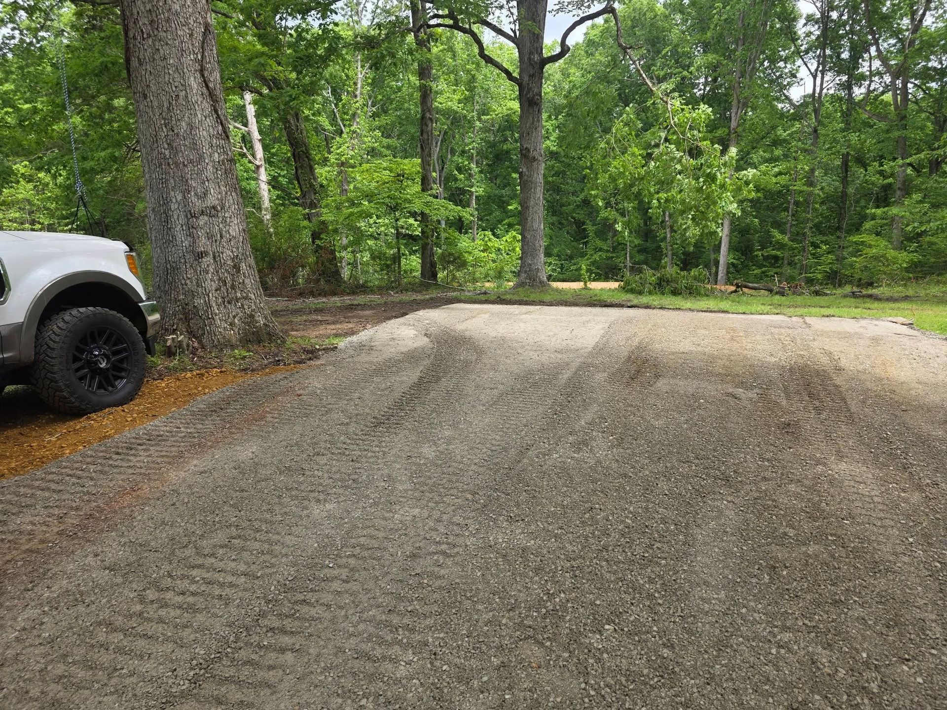 Gravel driveway next to a large tree, with a white truck parked on the left. Trees and greenery are in the background.