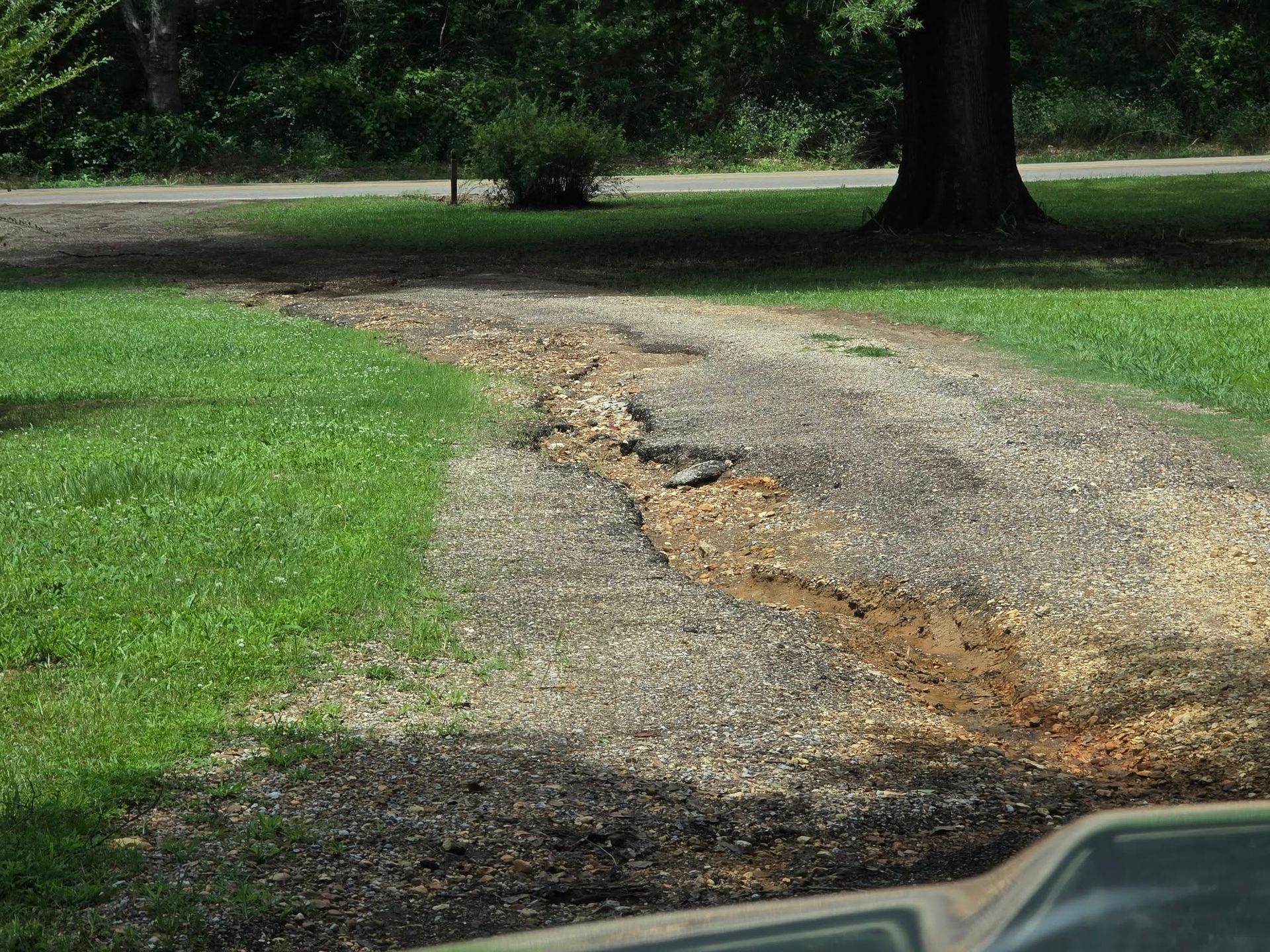 Gravel driveway eroded with water runoff, brown and gray, leading from grass to a road, outdoors.