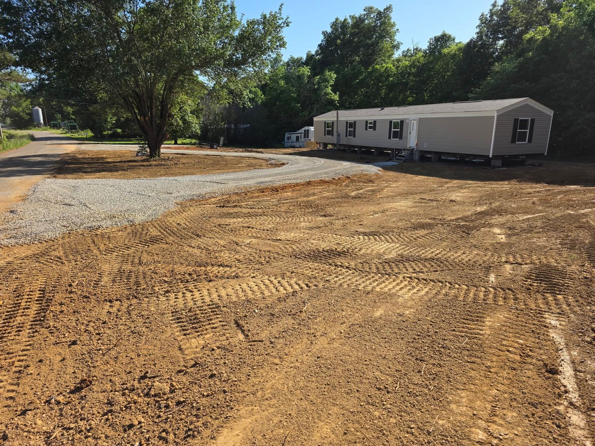 Mobile home on gravel lot, dirt foreground with tire tracks, tree on the left, sunny day.