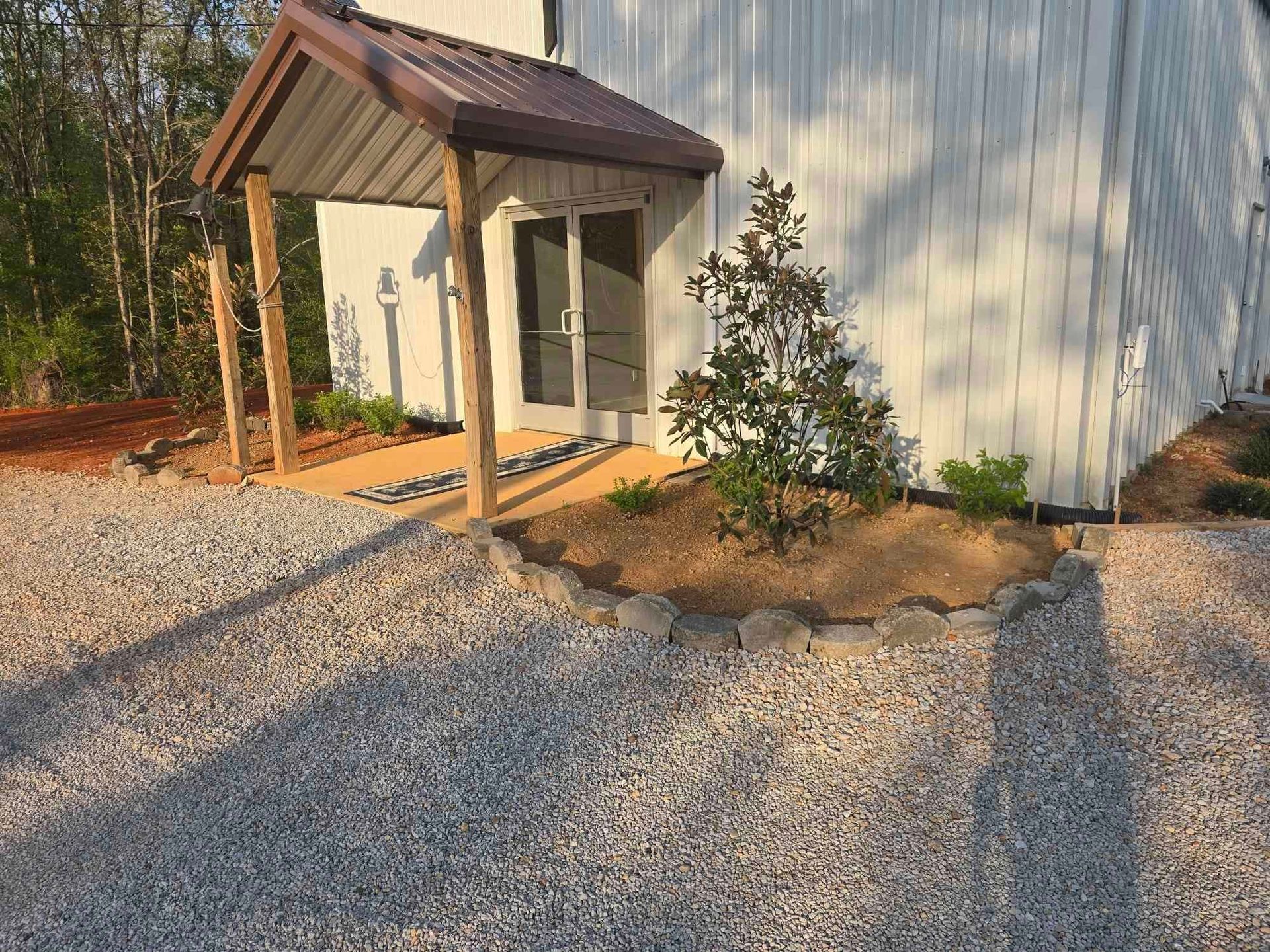 White building with gravel drive, brown awning over the door, a small tree in a raised bed.