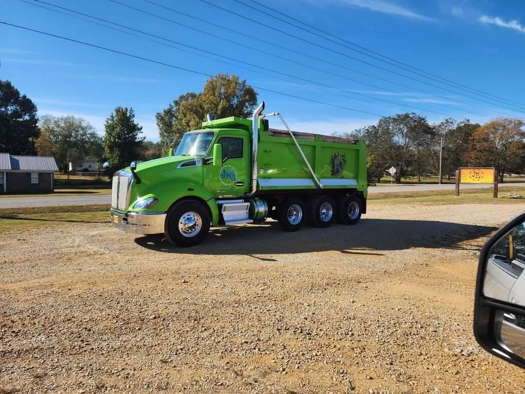Green dump truck parked on gravel lot under a blue sky.
