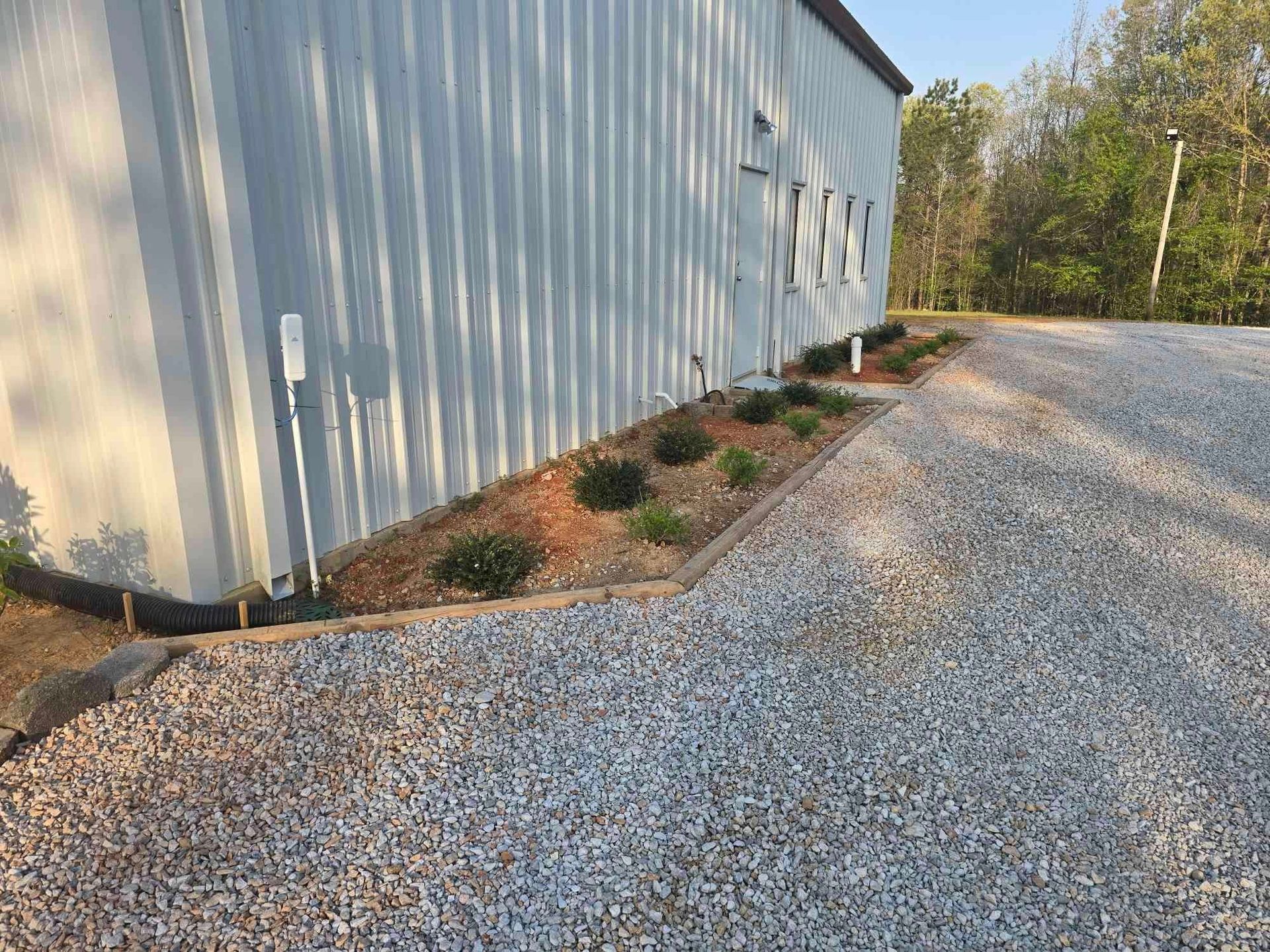 Gravel driveway next to a gray building with a small flowerbed containing green plants and brown mulch.