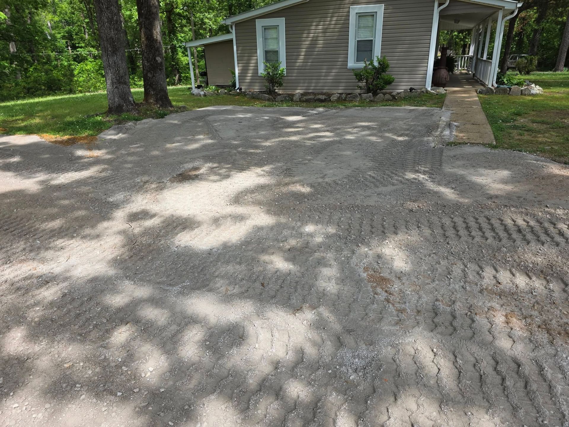 Gravel driveway in front of a light brown house, shaded by trees.