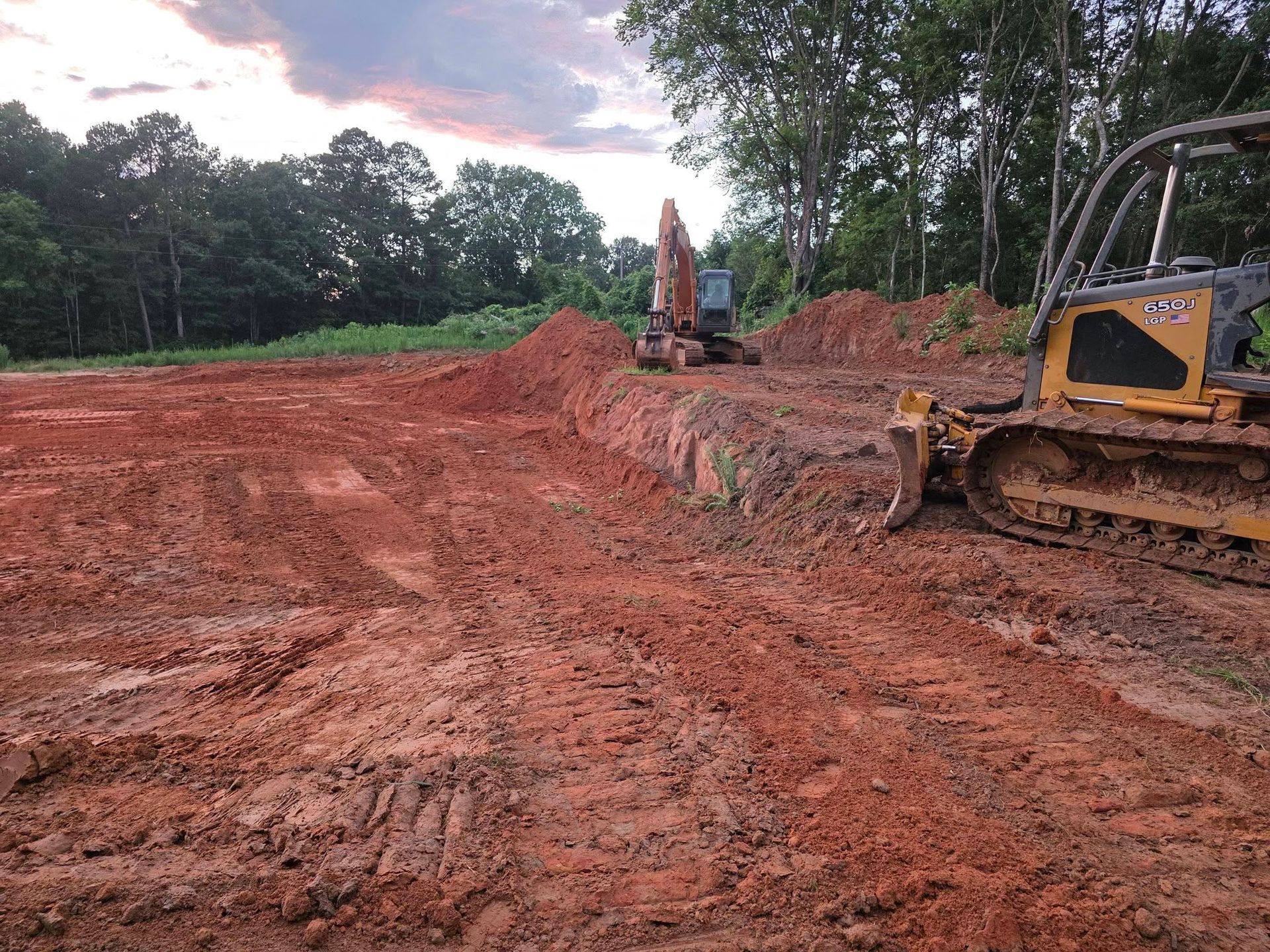 Construction site with red dirt, an excavator, and a bulldozer under a cloudy sky.