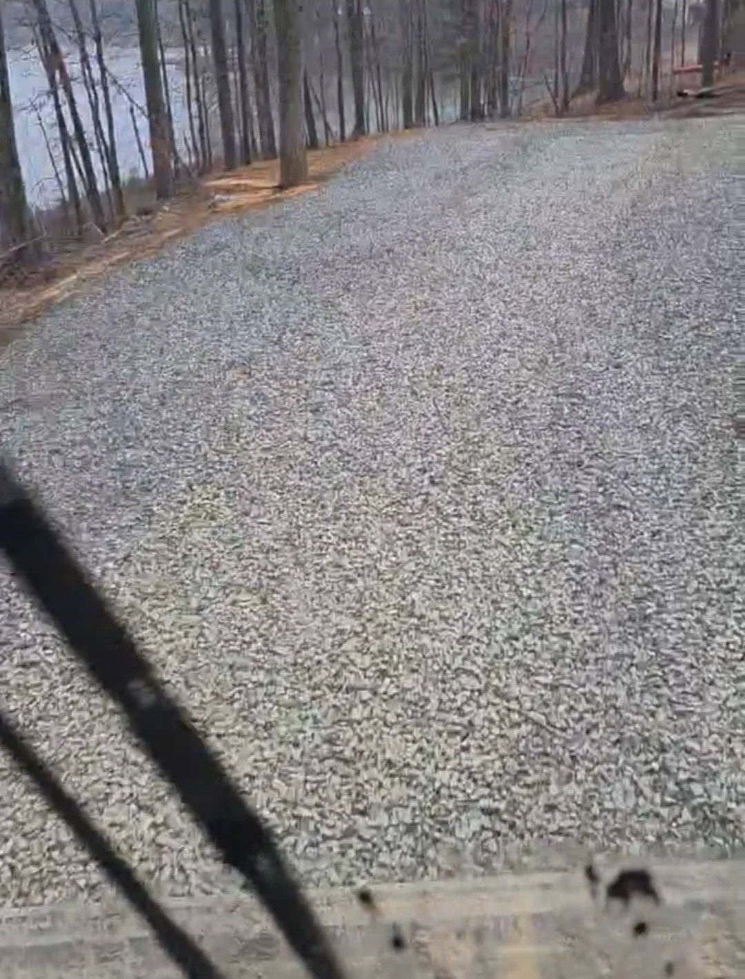 Gravel driveway leading uphill towards trees and a body of water in the distance.