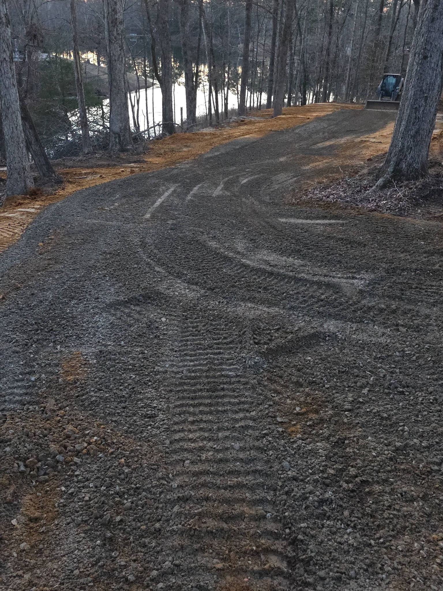 Gravel path through a wooded area leading towards a body of water. Tire tracks visible on the path.
