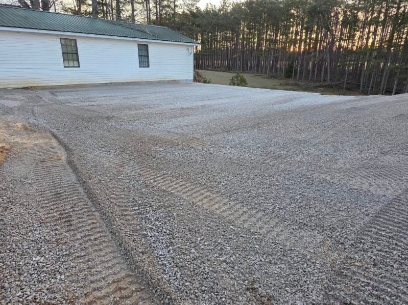Gravel driveway next to a white house with two windows; trees in the background.