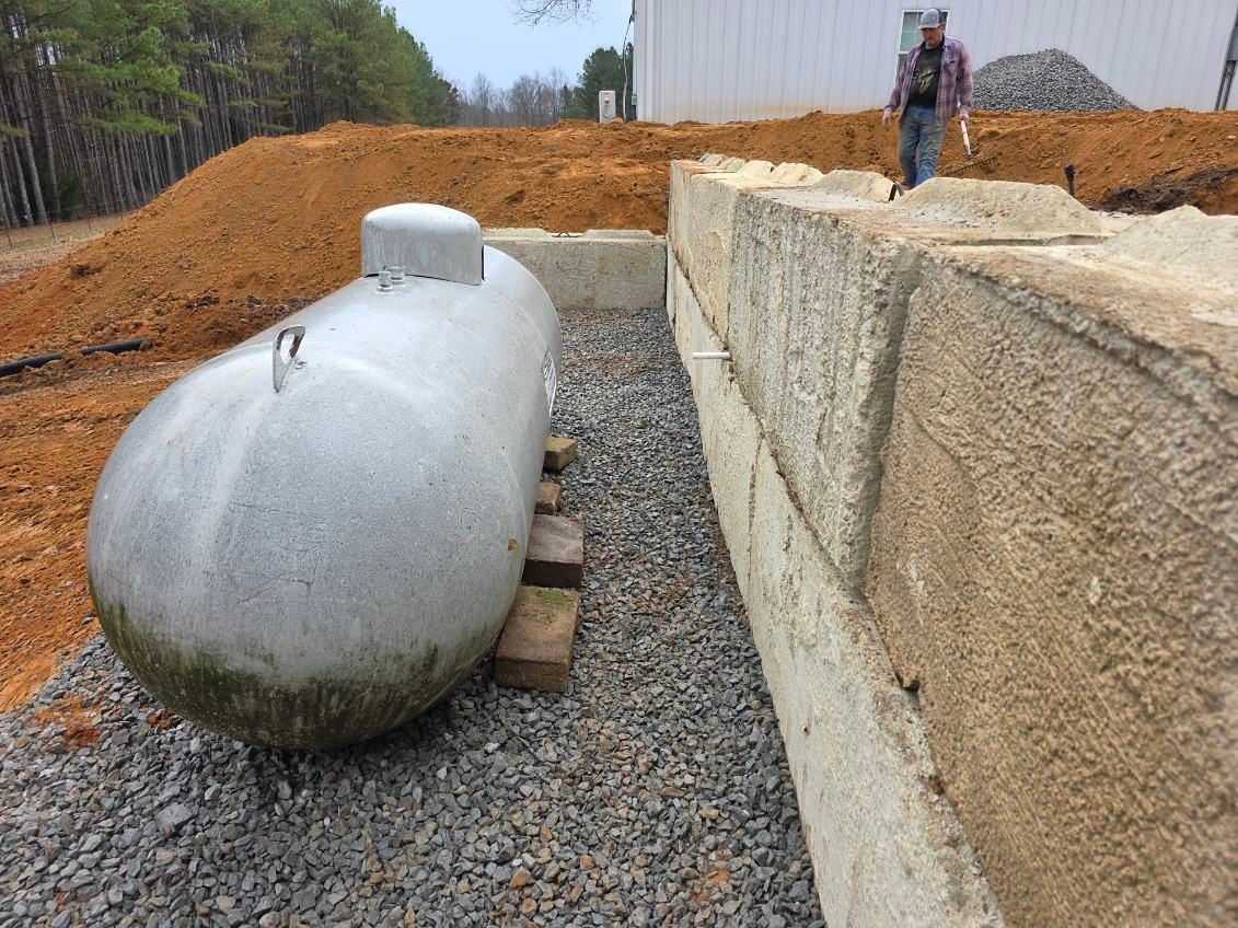 Large propane tank on gravel near a retaining wall, with a person standing on the wall, and dirt piles in the background.