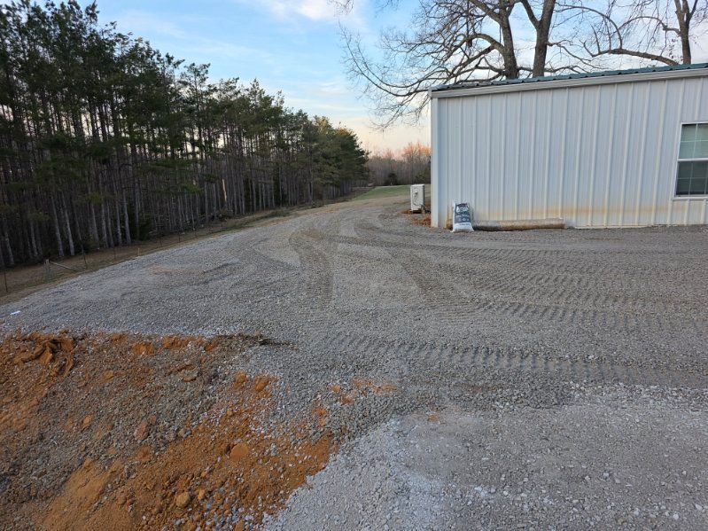 Gravel driveway extending from a small beige building to a road, surrounded by grass and blue sky.